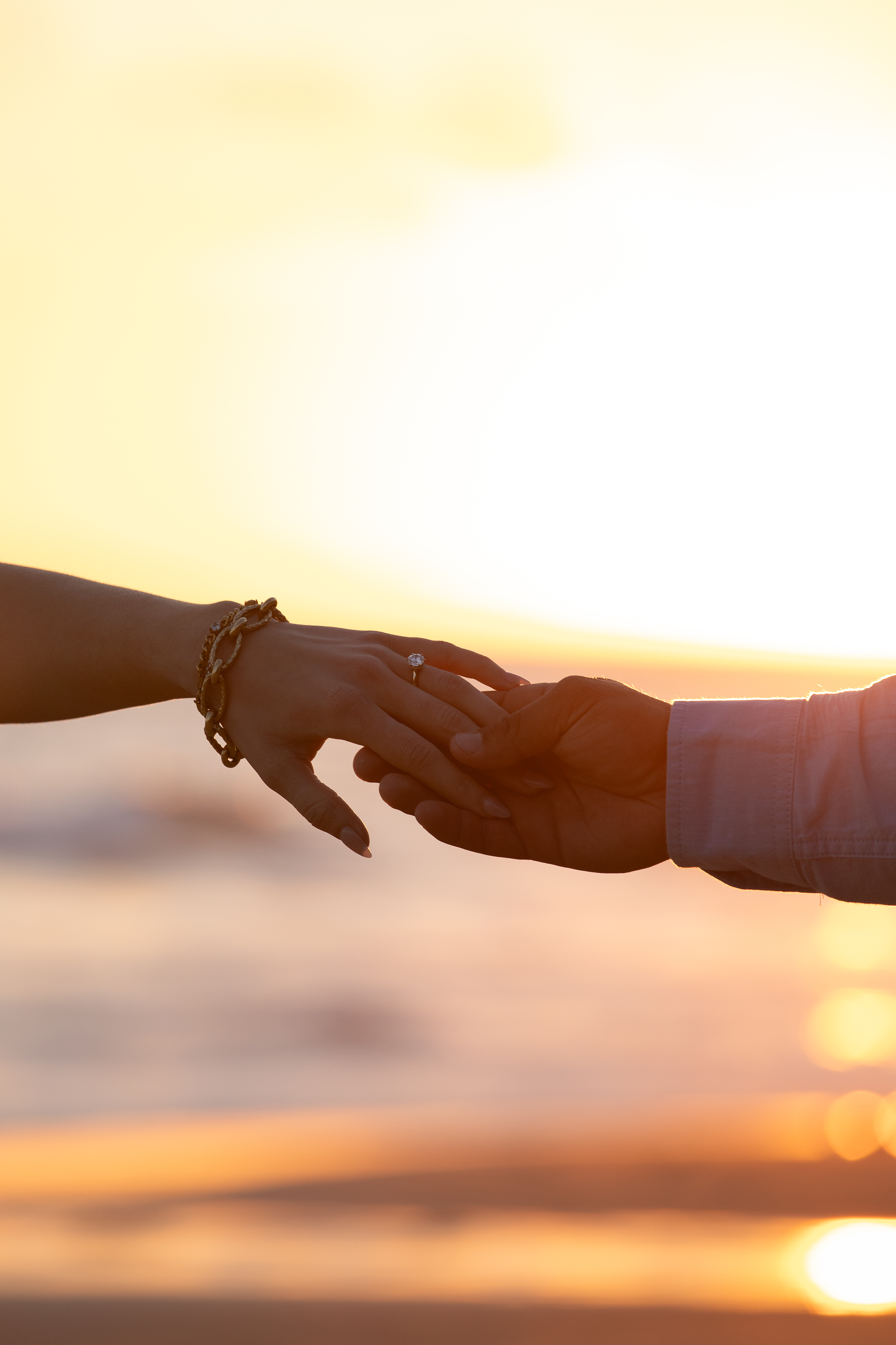 Silhouetted hands reaching for each other at sunset, engagement ring catching the light