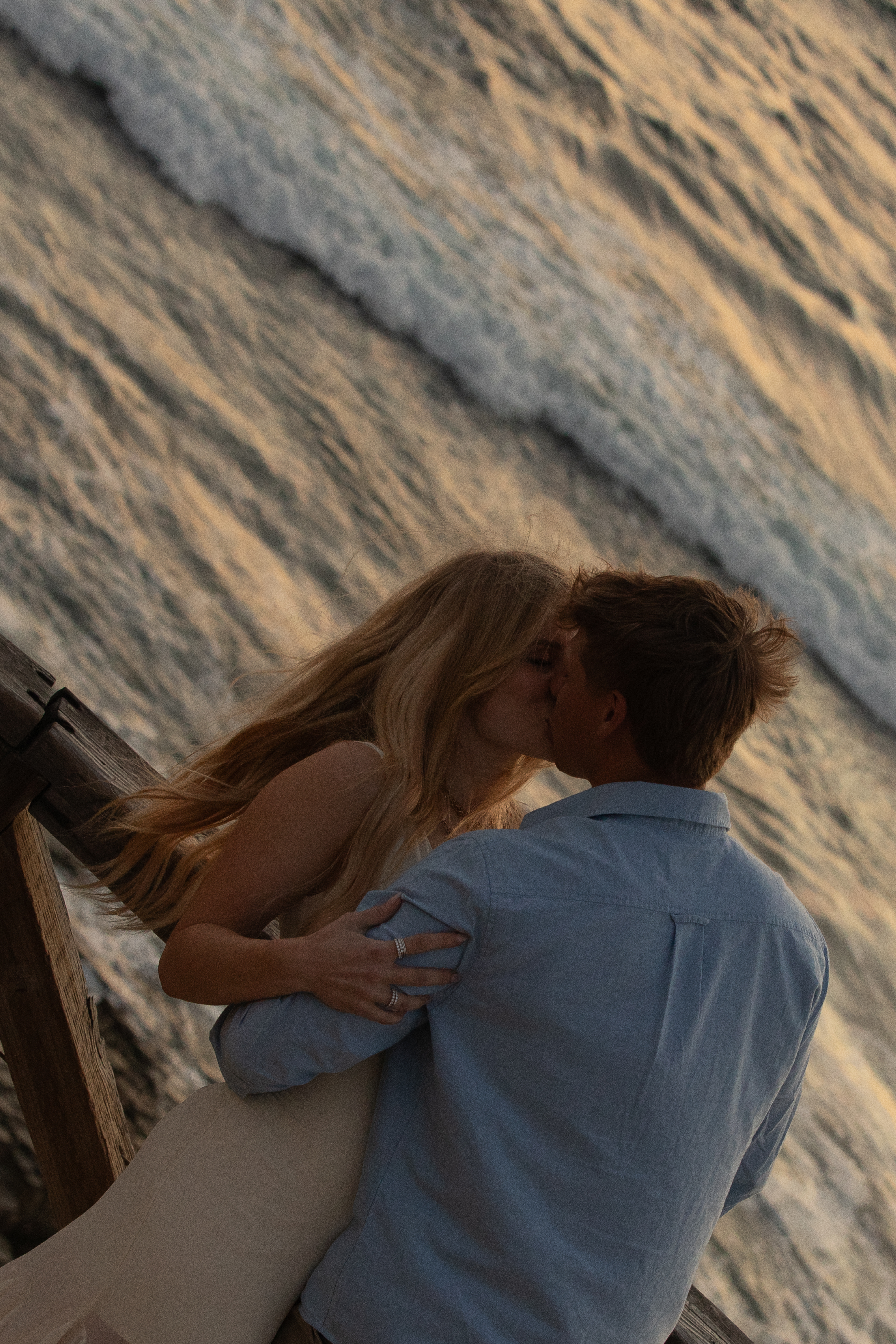 Engaged couple kissing on a weathered wooden pier with ocean waves behind them