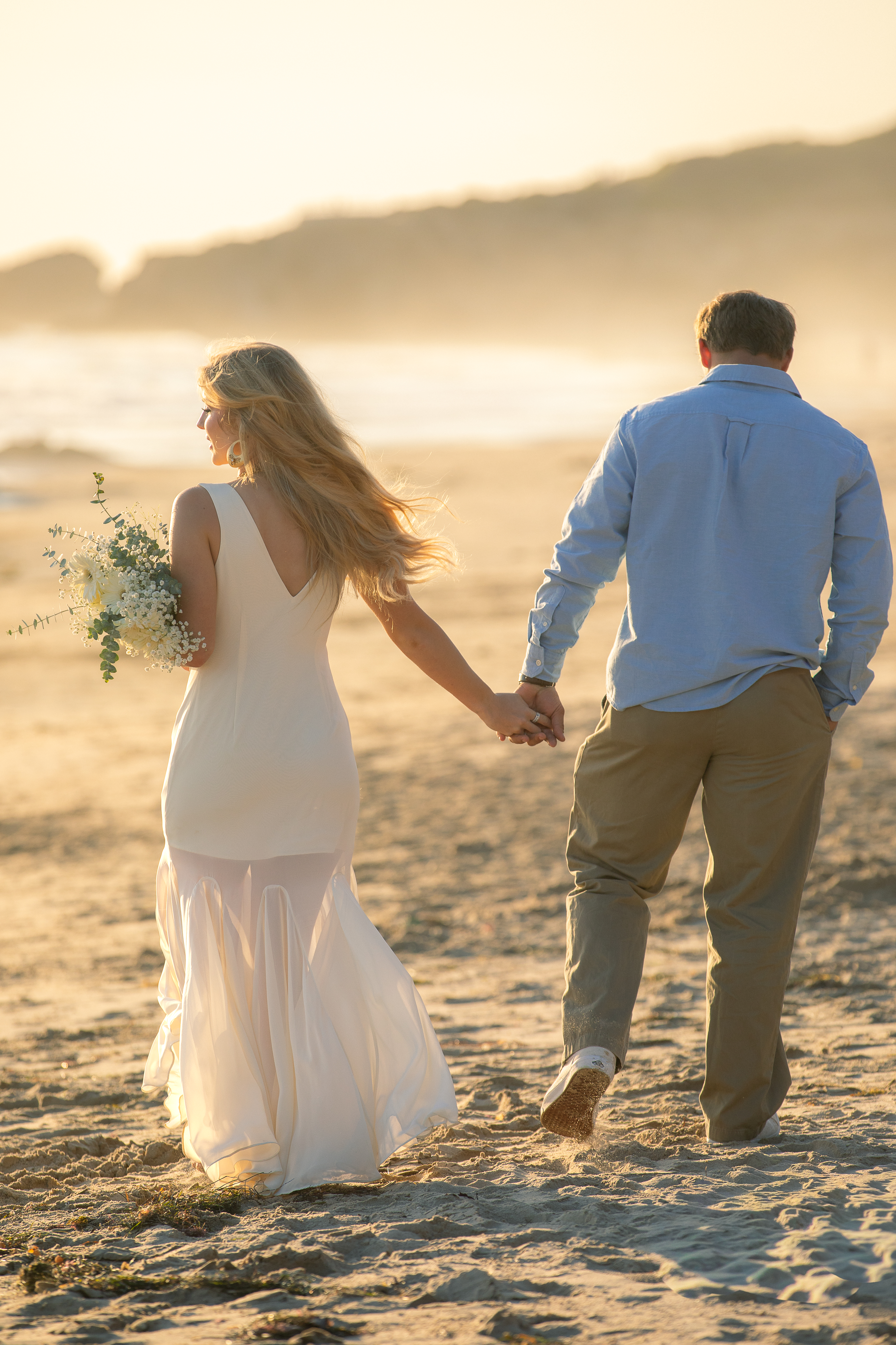 Engaged couple holding hands walking along the beach at sunset, bride holding a bouquet