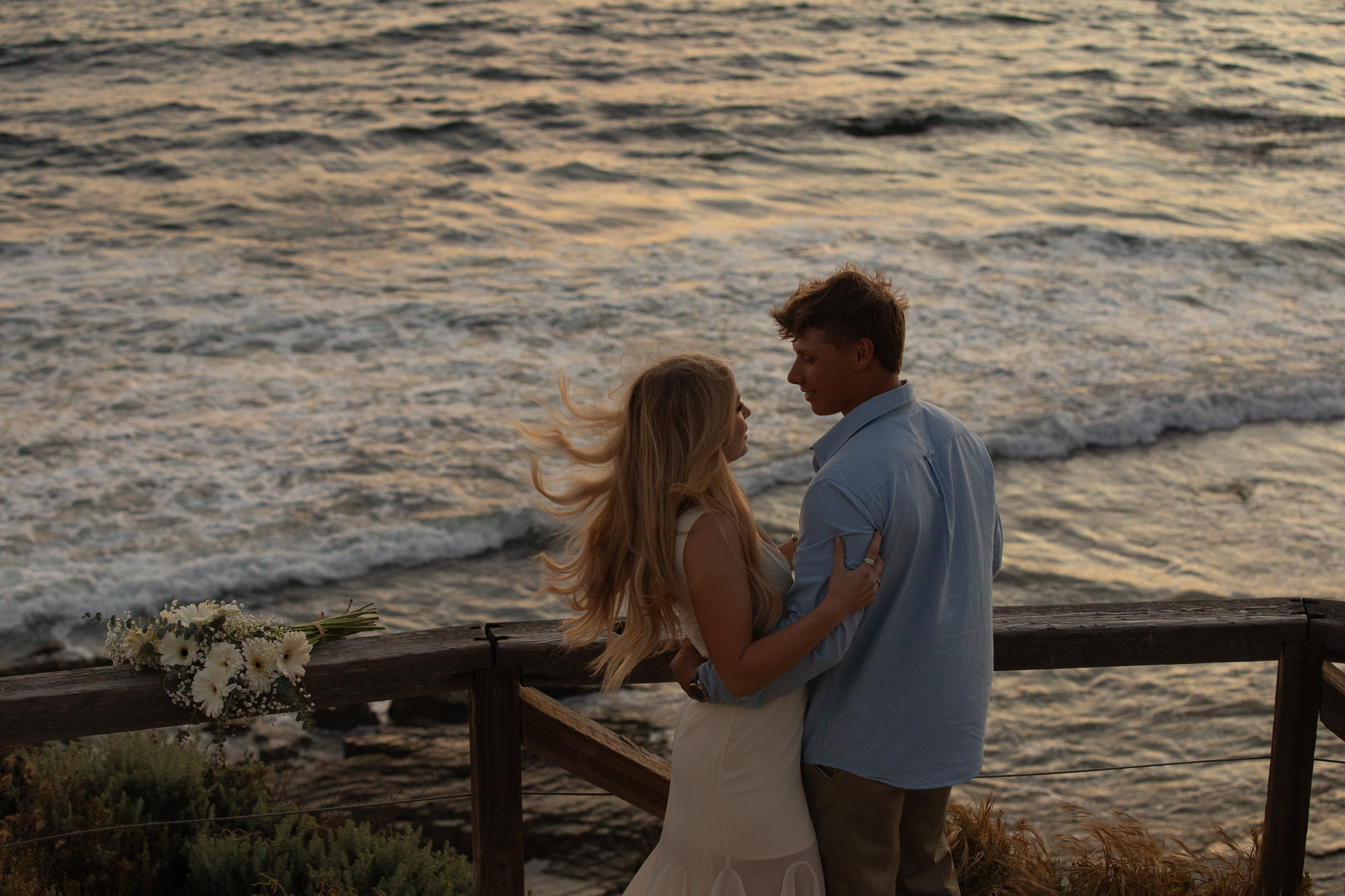 Couple holding each other on a wooden overlook above the ocean with a bouquet resting on the rail