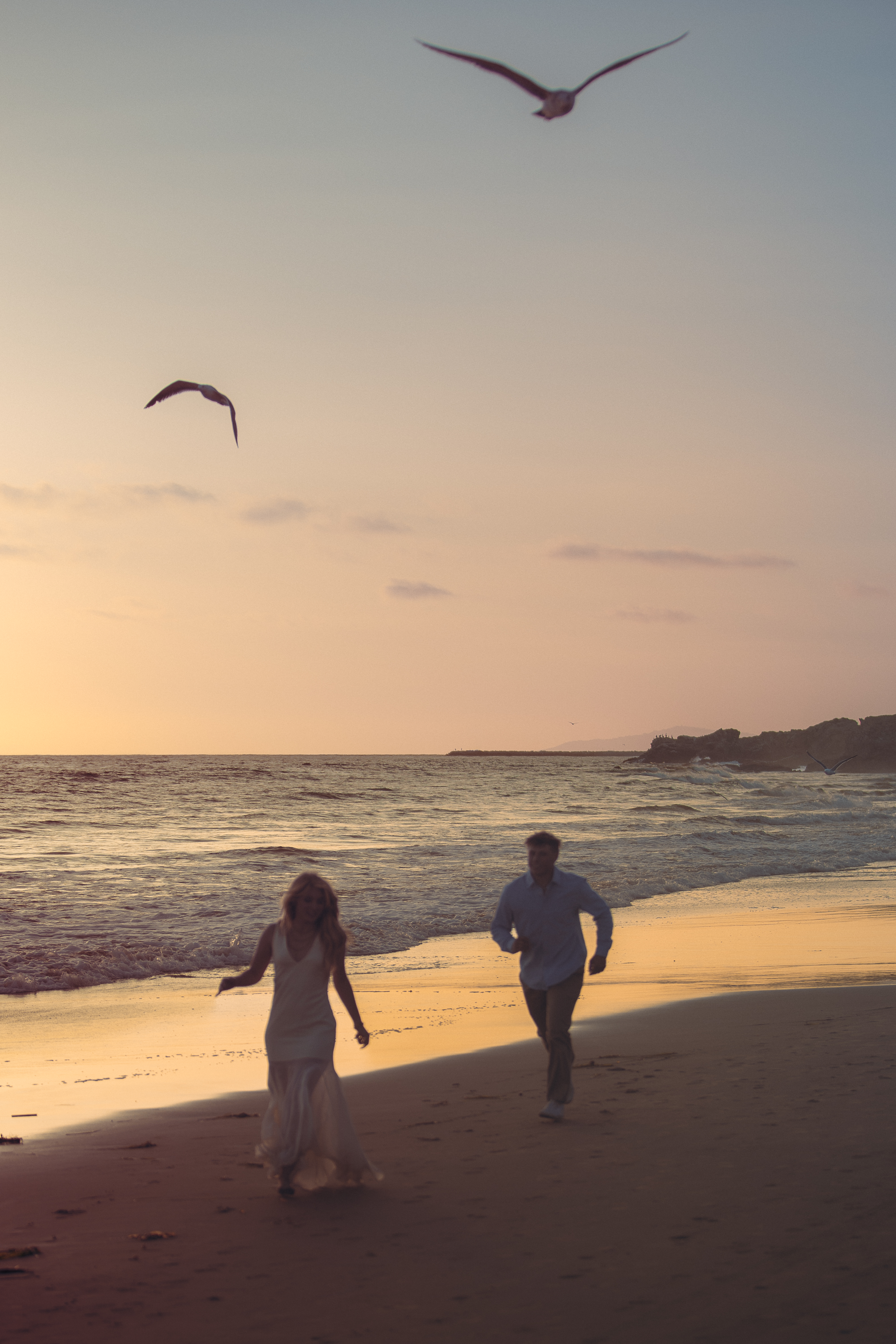 Couple running along the shoreline at sunset with seagulls overhead
