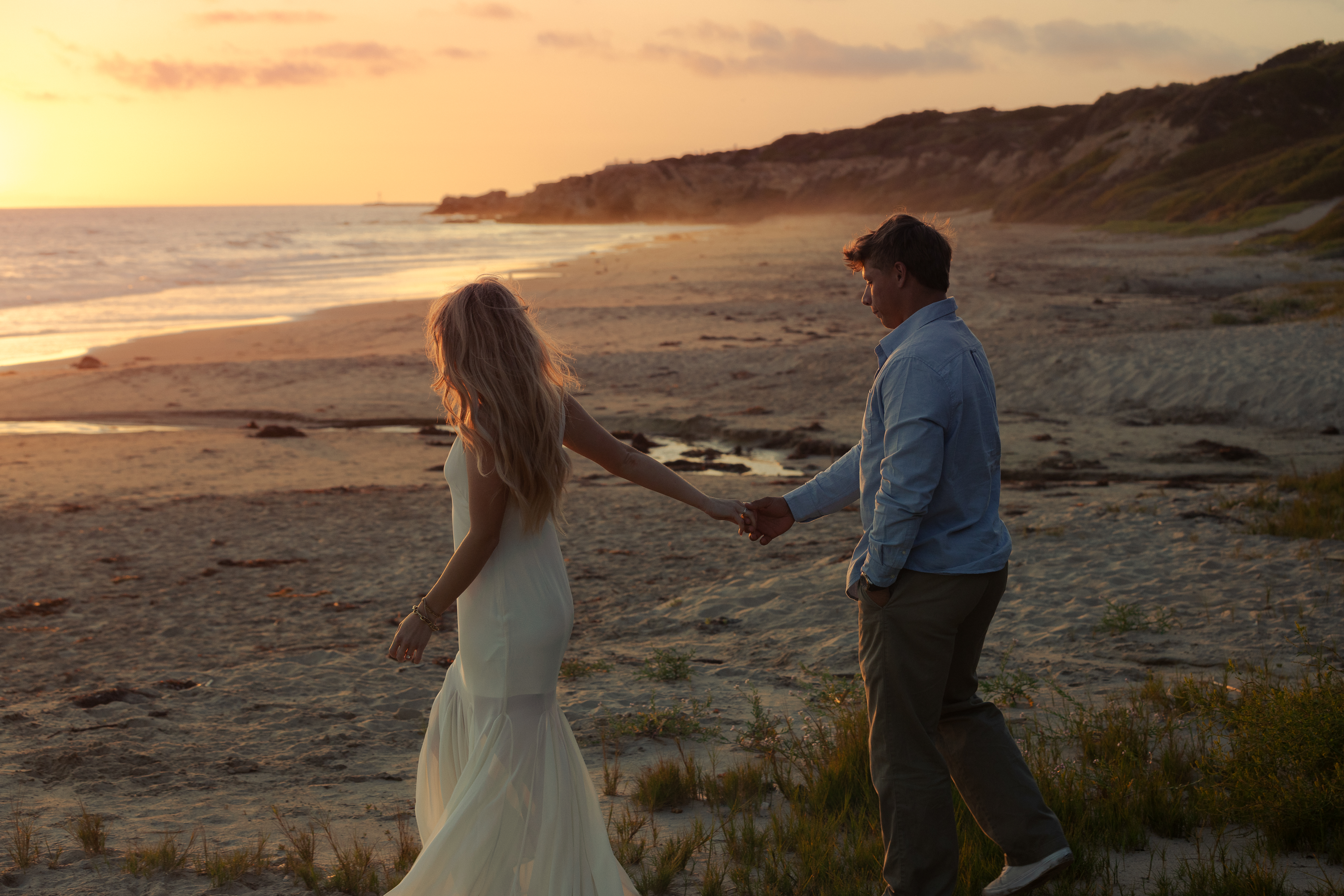 Couple walking hand in hand along the beach with coastal bluffs lit by sunset