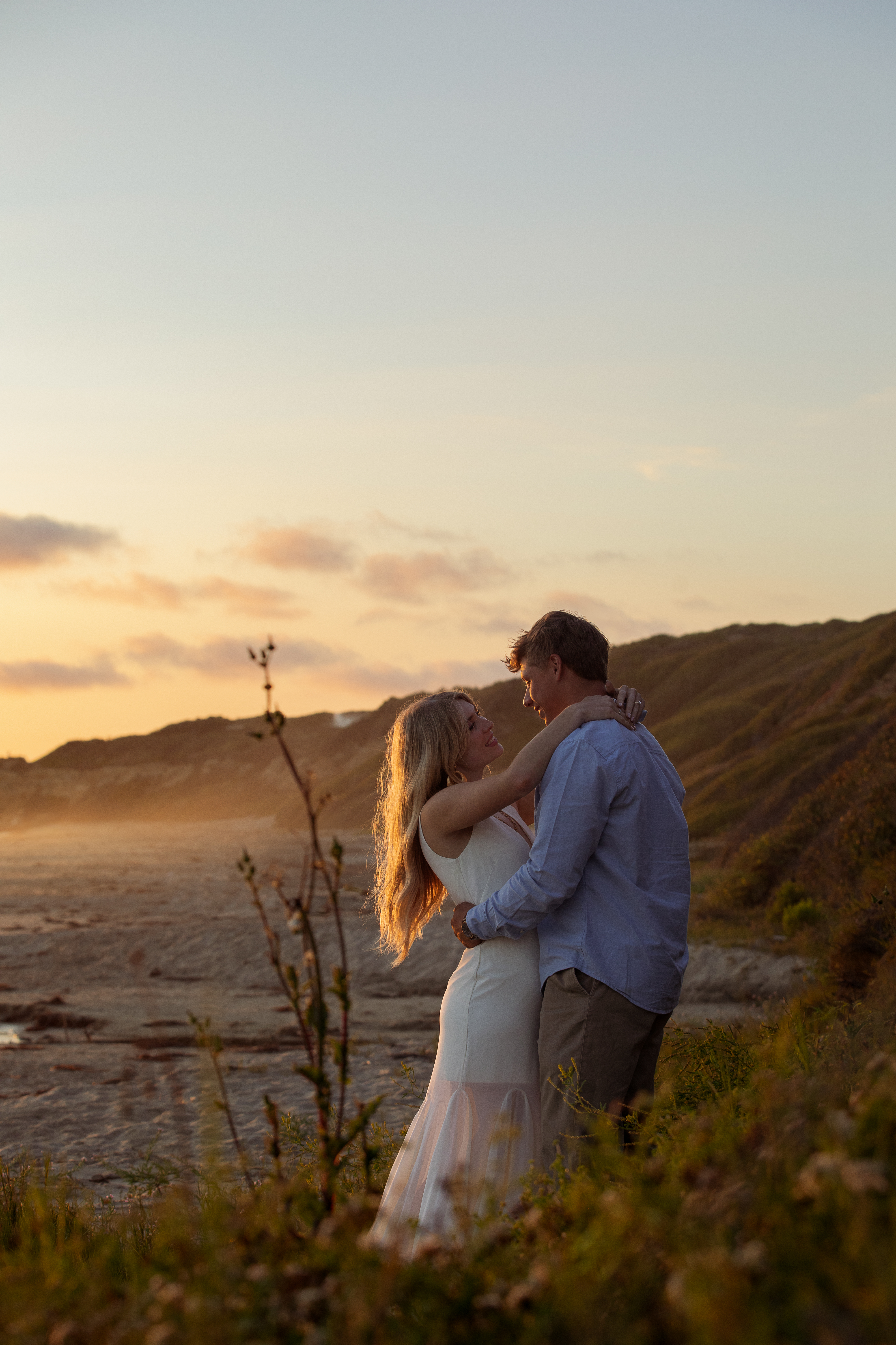 Couple embracing on a coastal bluff with hills and ocean behind them at sunset
