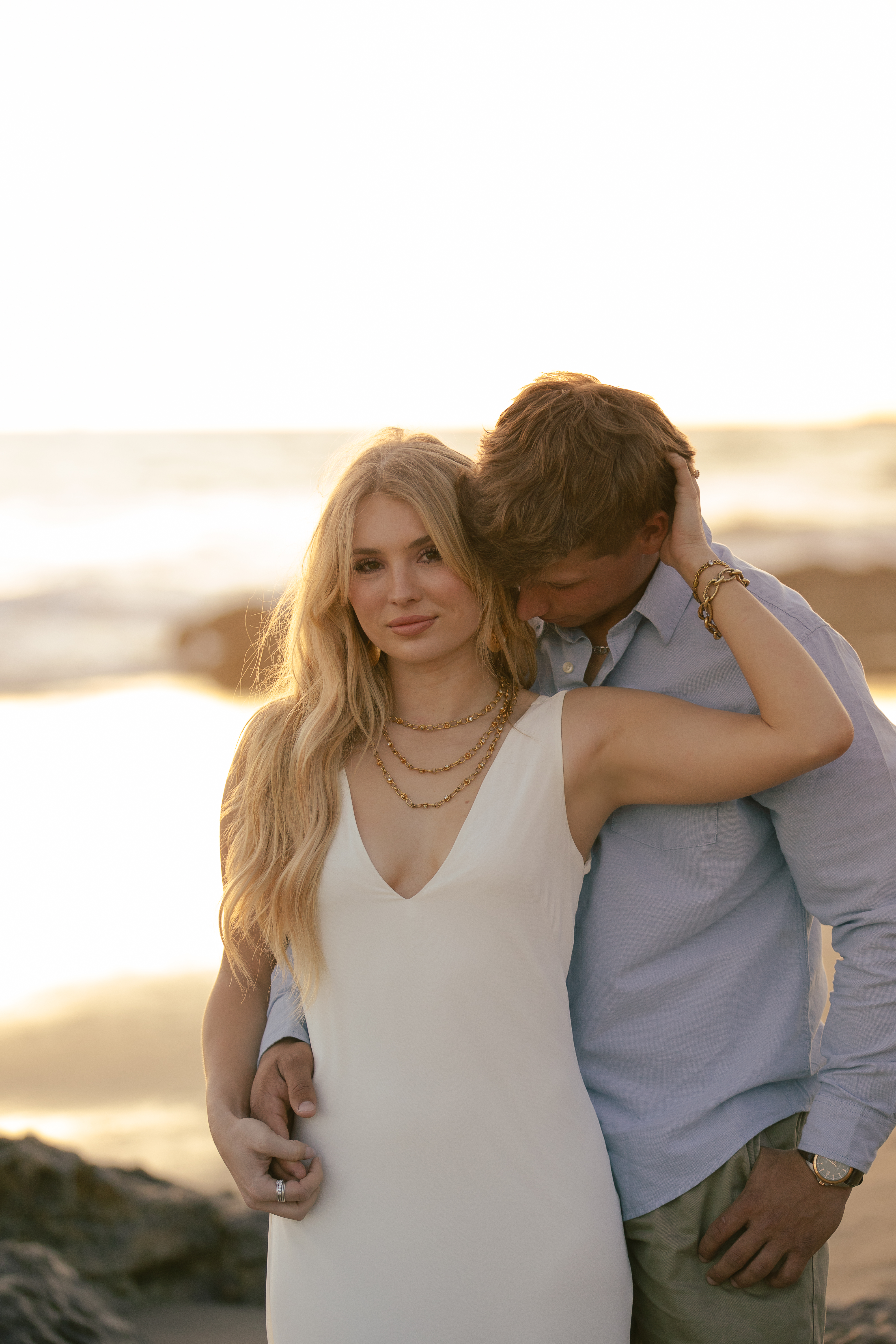 Engaged couple embracing at the beach at golden hour, groom resting his head on her shoulder