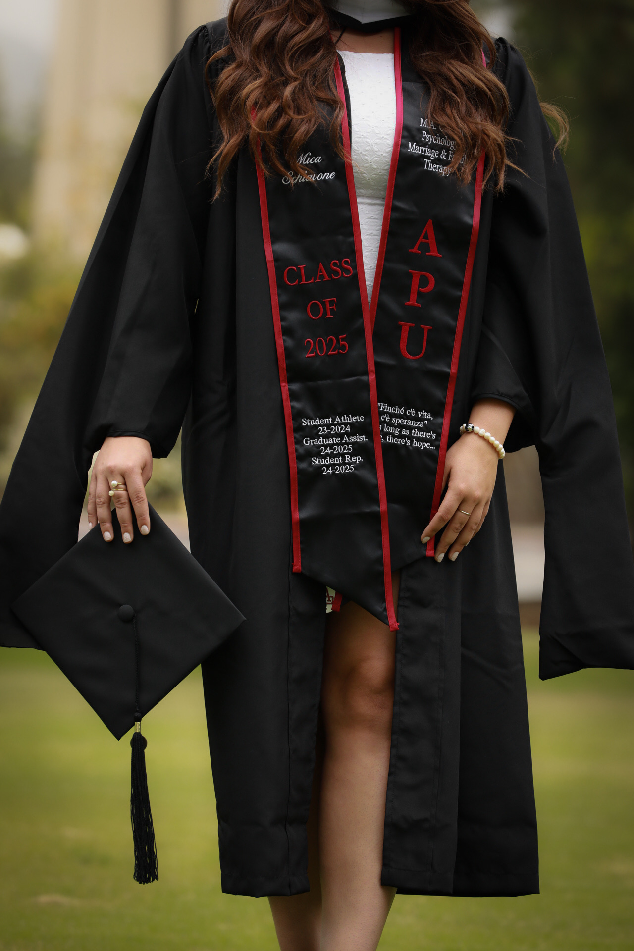 APU Class of 2025 graduate in a black gown holding her cap, detail shot of the red embroidered stole