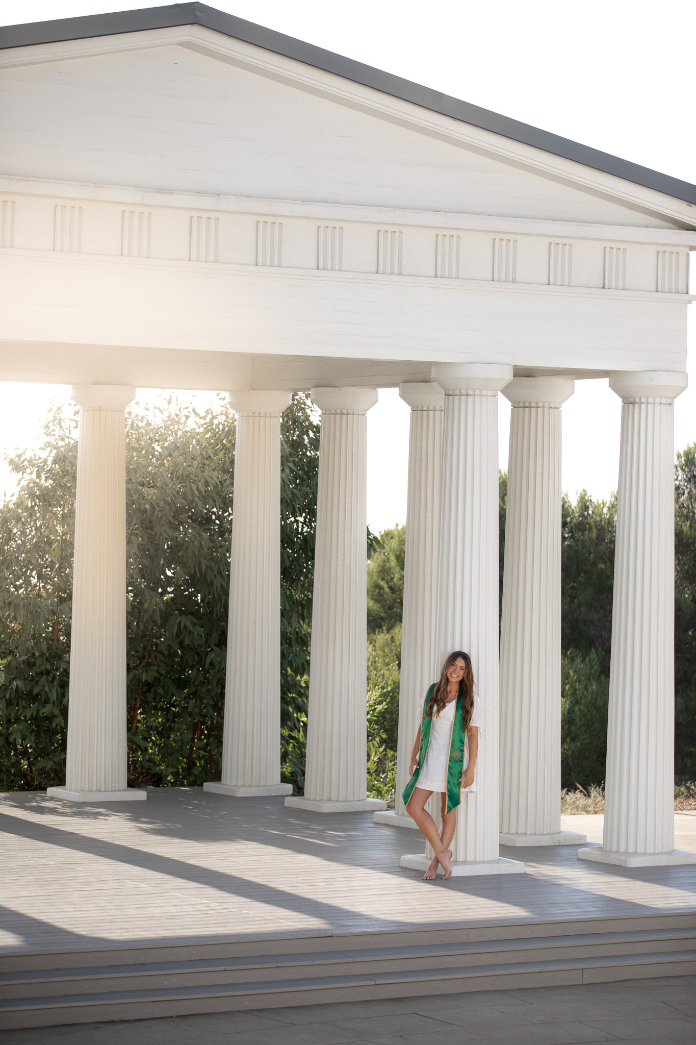 PLNU graduate in a white dress and green stole posing beneath a white colonnade