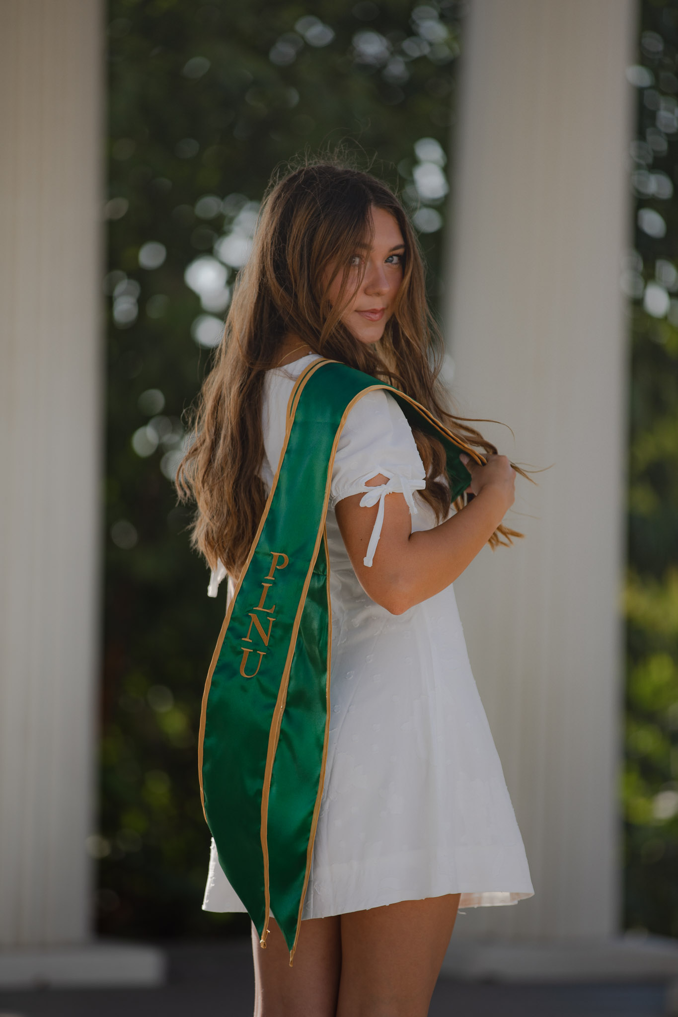 PLNU graduate in a white dress looking over her shoulder with her green graduation stole
