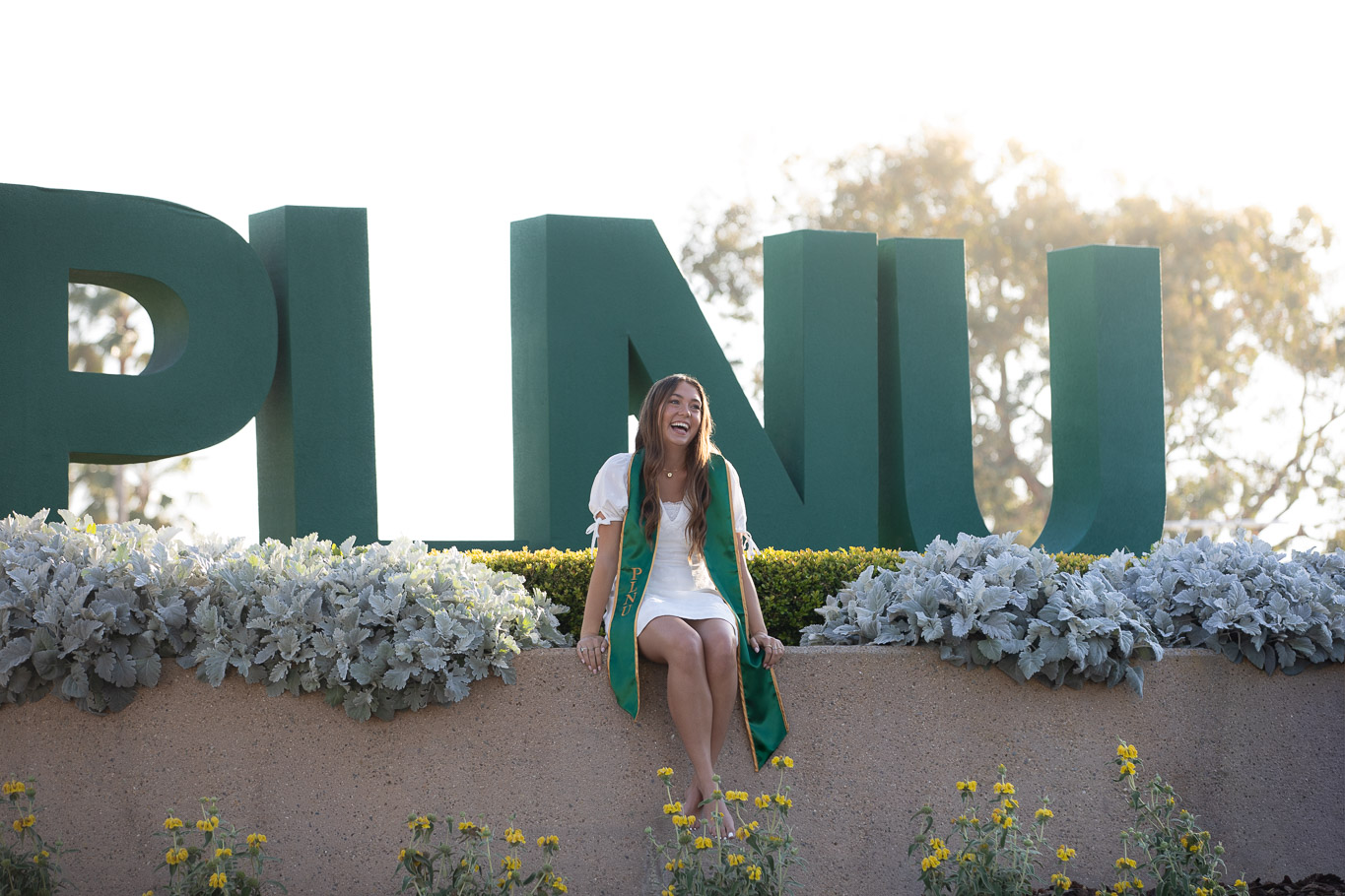 PLNU graduate laughing while seated in front of the campus PLNU letter sign