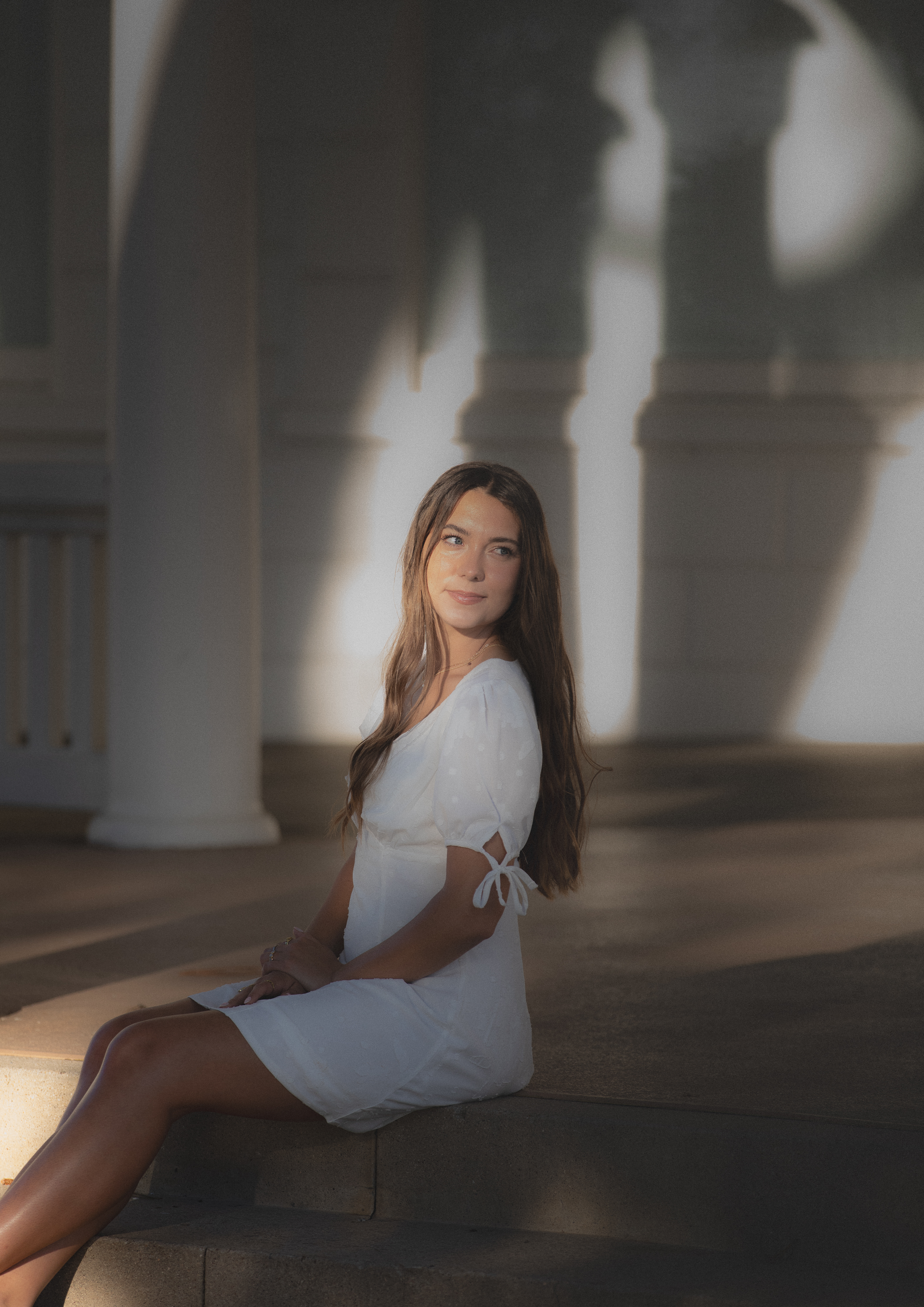 Senior graduate in a white dress seated on steps beneath an arched colonnade
