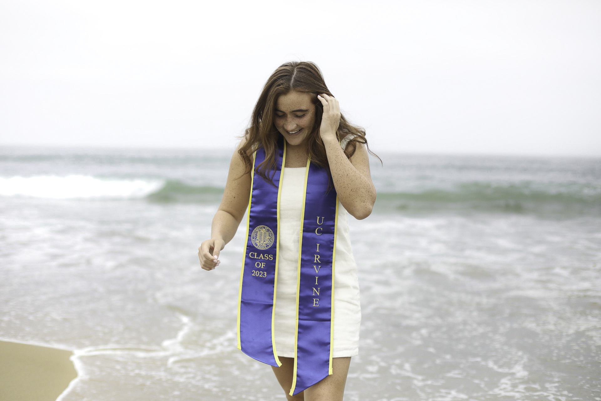UC Irvine Class of 2023 graduate smiling on the beach with waves behind her
