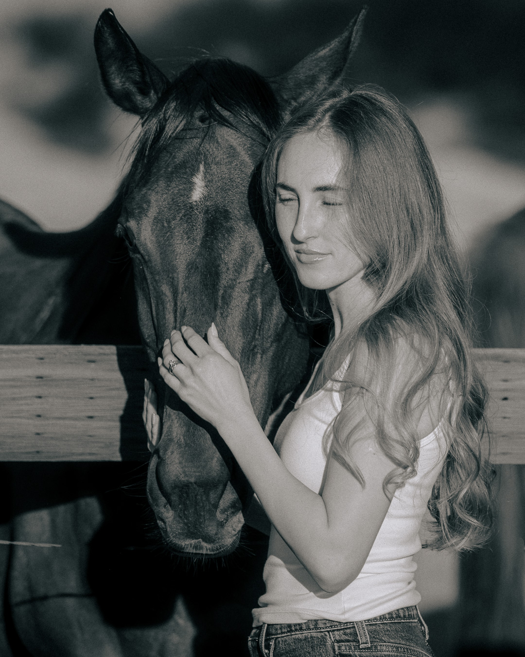 Black and white portrait of a woman resting her head against her horse at a wooden fence