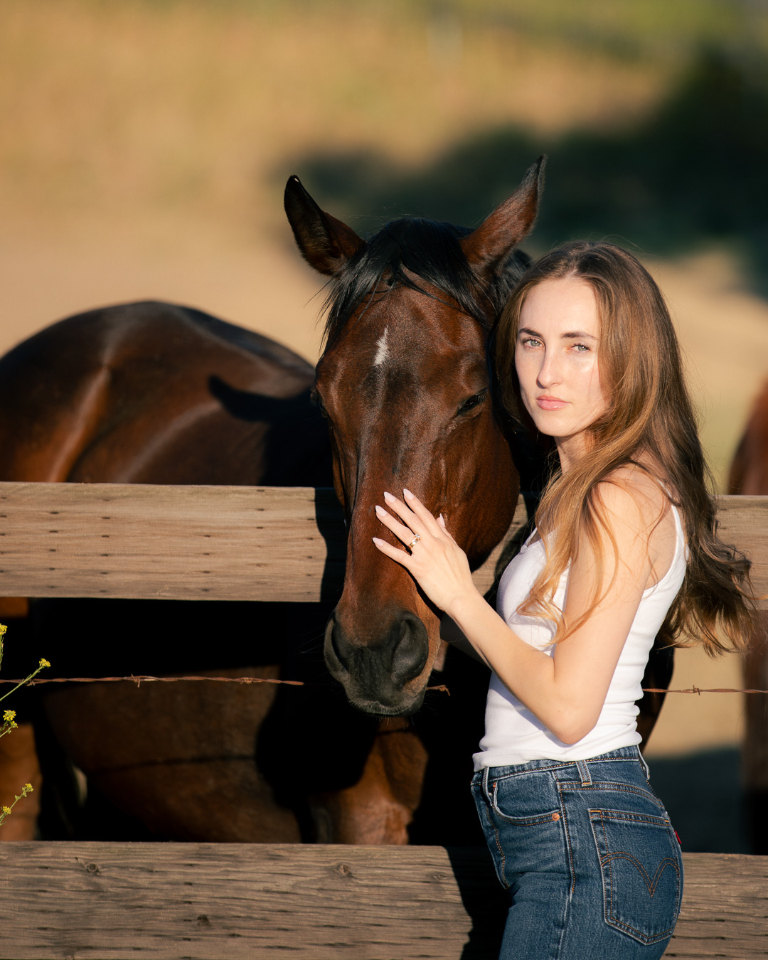 Woman in a white tank top and jeans standing beside her bay horse at a ranch fence