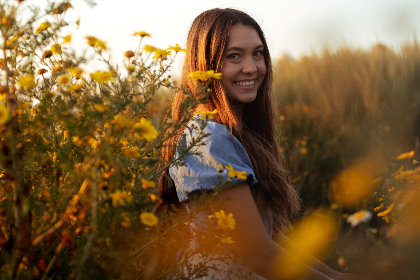 Close-up senior portrait of a smiling girl framed by yellow wildflowers in warm evening light