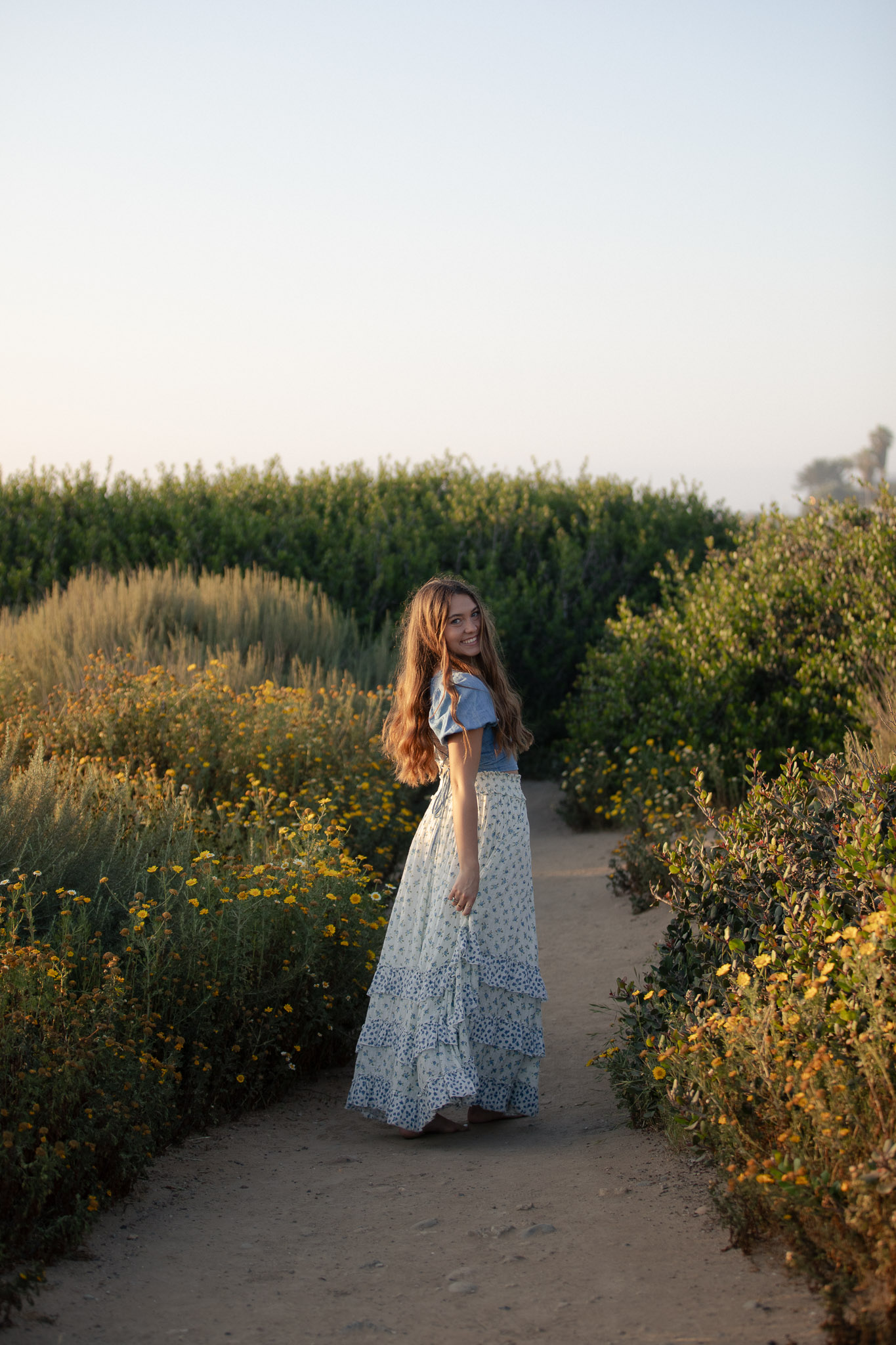 Senior portrait of a girl in a flowing floral skirt on a sandy path lined with wildflowers