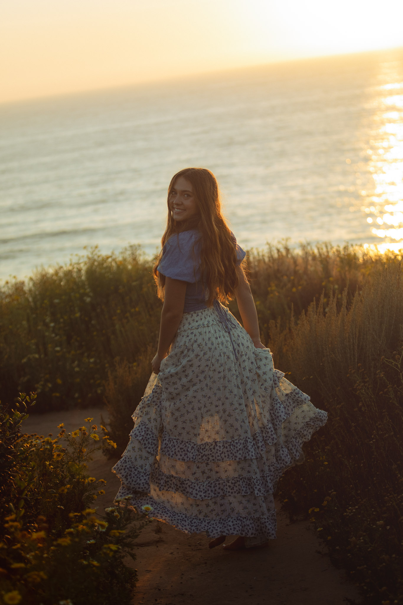 Senior portrait of a girl in a floral skirt twirling on a coastal bluff at sunset